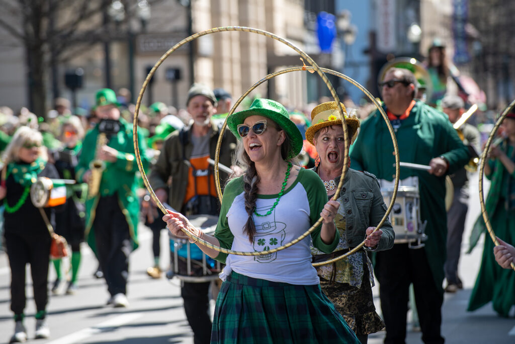 A member of the March Madness Marching Band celebrates during the Lexington St. Patrick’s Parade, wearing a green sequined hat and holding a gold hula hoop as fellow band members perform in the background.