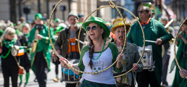 A member of the March Madness Marching Band celebrates during the Lexington St. Patrick’s Parade, wearing a green sequined hat and holding a gold hula hoop as fellow band members perform in the background.